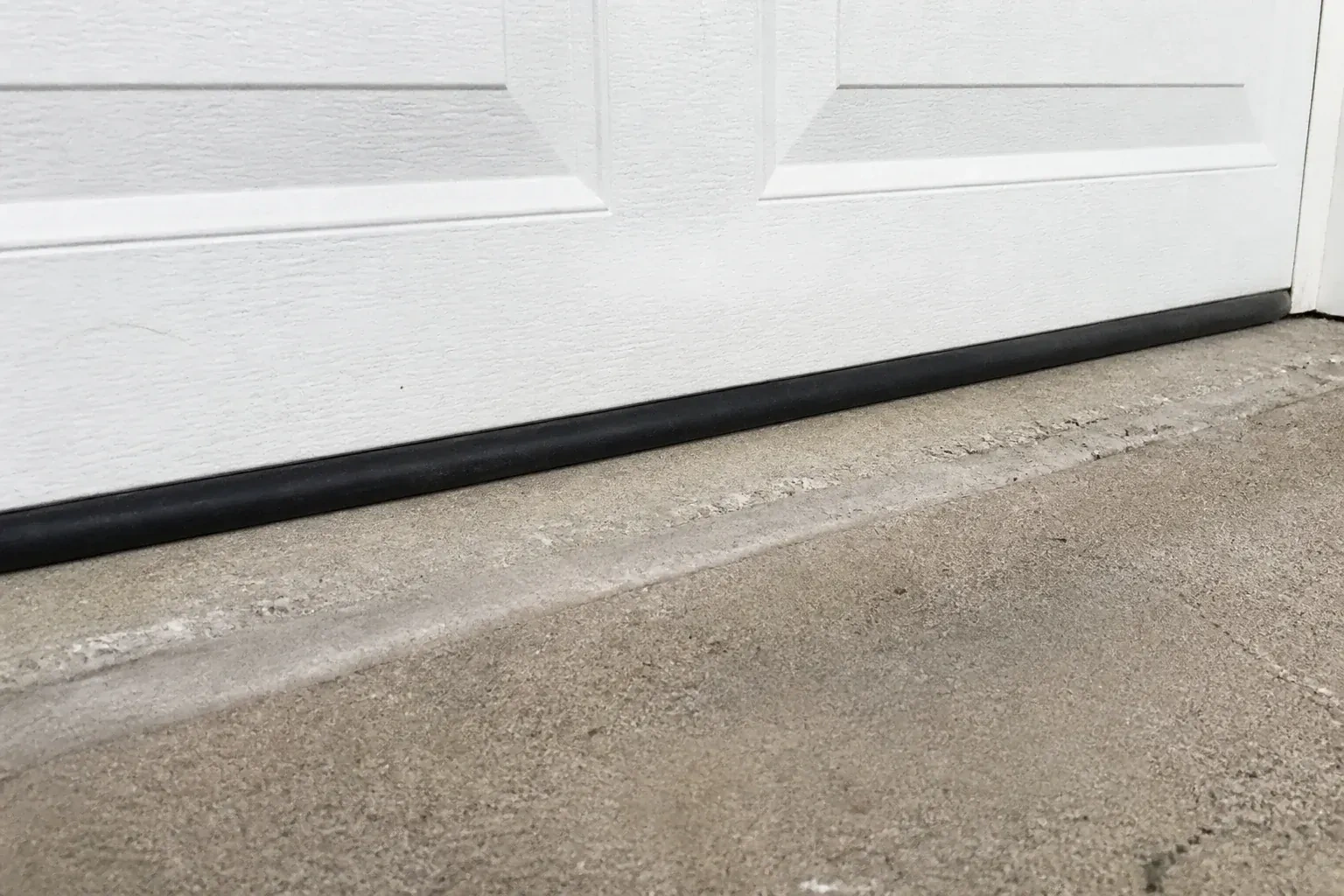 Black bottom seal along a closed white garage door at a concrete threshold. — Garage Door Bottom Seal Replacement, Oshawa